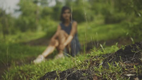 Close-up-fixed-shot-of-earth-and-grass-seedlings-on-the-side-of-a-gentle-hill,-with-female-south-asian-model-sitting-in-the-grass-blurred-in-the-background