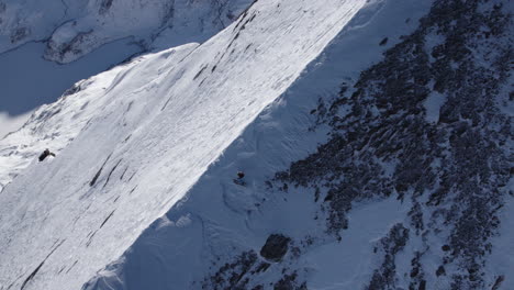 Aerial-tracking-shot-of-skier-carving-down-snowy-steep-mountain-during-sunlight---Spectacular-drone-shot-of-professional-skier-on-rocky-scarp