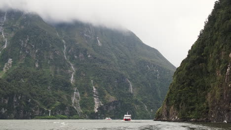 Ein-Boot-Fährt-An-Einer-Hoch-Aufragenden-Klippe-Entlang,-Während-Im-Hintergrund-Ein-Majestätischer-Wasserfall-Stürzt