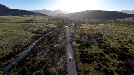 Aerial-shot-of-car-driving-on-dirt-road-in-Willcox,-Arizona,-wide-drone-shot-with-mountains-in-the-background-and-dust-behind-the-car-into-the-sun