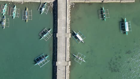 Aerial-Top-Down-View-of-multiple-Filipino-Outrigger-Bangka-Boats-moored-to-a-quaint-dock,-revealing-tropical-village-town-and-settlers-inland