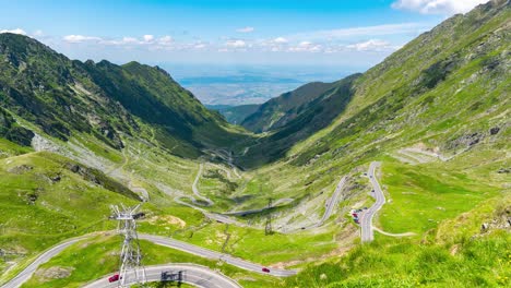 Transfagarasan-Highway-A,-Timelapse-Of-Towering-Peaks,-Lush-Green-Valleys,-and-Clear-Blue-Skies-with-Fast-Moving-Clouds,-Red-Cable-Cars,-and-Cars-Driving-up-and-down-the-Mountain