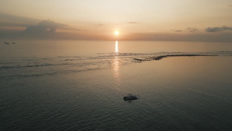 boat-drifting-during-Bali-sunrise,-cinematic-aerial,-the-sun-is-rising-on-the-horizon-of-a-calm-sea