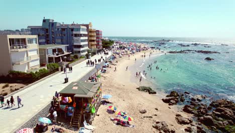 Fly-over-a-busy-beach-with-a-lifeguard-store-and-umbrellas-to-protect-from-the-sun,-sunny-day-at-Pejerrey-beach-in-Algarrobo