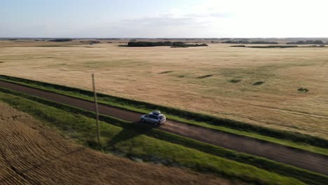 Aerial-tracking-shot-of-silver-car-driving-along-dirt-road-in-the-countryside-of-Alberta,-Canada