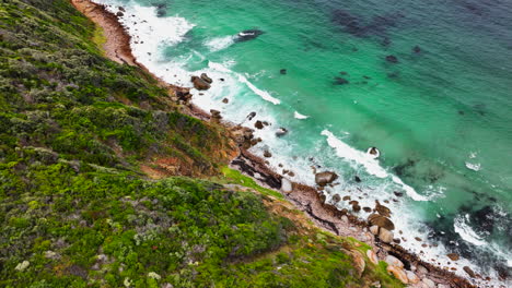 South-Africa-small-beach-town-Smitswinkel-Bay-Cape-of-Good-Hope-Simon's-Town-aerial-drone-cinematic-homes-surf-wave-green-lush-spring-summer-deep-aqua-blue-ocean-cloudy-mountain-pan-down-movement
