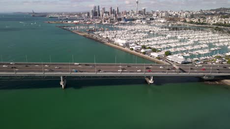 Traffic-on-Auckland-Harbour-Bridge,-aerial-view-of-Royal-Yacht-Squadron-Club-and-cityscape-with-Sky-Tower