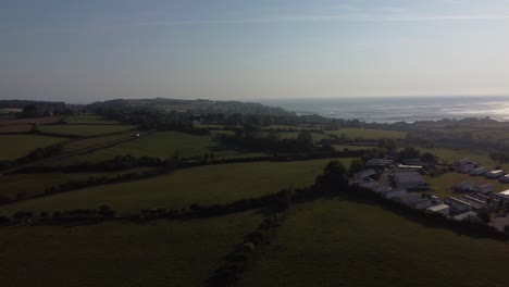 Aerial-view-Welsh-caravan-park-at-sunrise-overlooking-shimmering-coastal-bay-and-rolling-countryside-hills,-panning-right