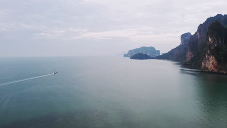 Boat-cruising-across-sea-along-mountainous-coastal-cliffs-in-Thailand