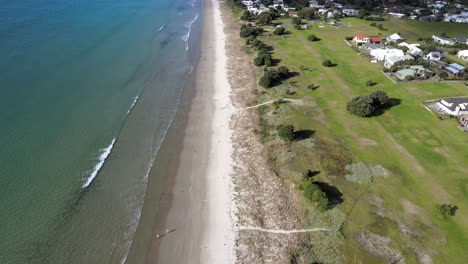 Matarangi-beachfront-aerial-tilt-up-from-sandy-beach-to-New-Zealand-coastline,-sunny-day