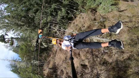 Vertical-action-camera-shot-of-a-young-man-swinging-by-very-close-to-trees-and-bushes-in-the-Canadian-forest-on-the-zipline-in-the-hard-summer-sun-on-his-holiday