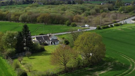 Rural-British-farmhouse-aerial-view-surrounded-by-lush-green-trees-and-agricultural-farmland-countryside-fields,-Orbit-right-shot
