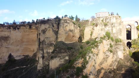 Ronda-cityscape-on-top-of-steep-rocky-cliff-on-sunny-day,-aerial-side-fly-view