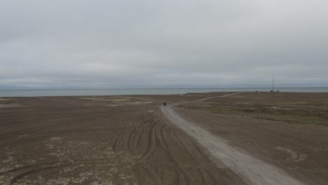 Aerial-Tracking-Drone-Shot-of-Solitary-Man-on-ATV-on-Black-Sand-Beach-and-Arctic-Ocean-at-the-Northernmost-Point-of-the-Arctic-United-States-near-Barrow-Alaska