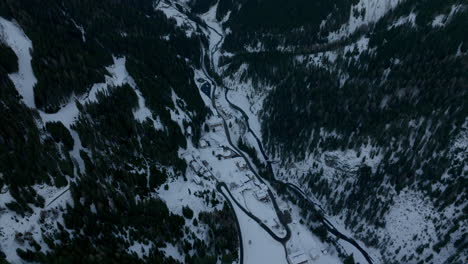 Flyover-Drone-Shot-of-winding-roads-of-Stelvio-Pass-alongside-river-in-between-dense,-coniferous-forest