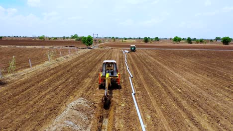 Aerial-camera-moving-forward-to-digging-soil-at-agricultural-land-on-daytime,-excavation-working-and-digging-for-water-pipeline-at-agricultural-farm