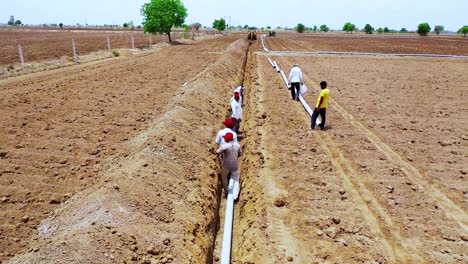 Indischer-Bauer-Und-Arbeiter-Arbeiten-In-Heißen-Sommermonaten-An-Der-Installation-Einer-Wasserableitungsleitung,-Luftkamera-Folgt-Dem-Arbeiter