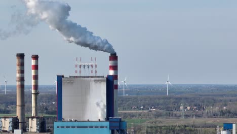 aerial-of-power-plant-chimney-release-flue-gas-smoke-into-the-atmosphere-with-wind-turbine-in-the-background,-green-energy-pollution-zero-emission-concept