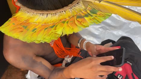 Ein-Indigener-Amazonischer-Mann,-Gekleidet-In-Einen-Wunderschönen-Kopfschmuck-Mit-Bunten-Federn,-Schaut-Auf-Ein-Smartphone