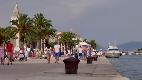 People-stroll-along-the-marina-in-Trogir,-Croatia,-with-boats-and-palm-trees-in-this-static-shot