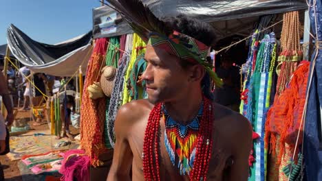 An-Amazon-indigenous-man,-traditionally-dressed-in-beaded-pendants-and-a-feathered-hat,-stands-in-front-of-his-market-stall