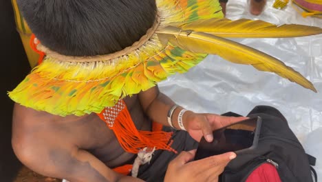 Ein-Indigener-Mann-Aus-Amazonien,-Gekleidet-In-Einen-Farbenfrohen-Federhut,-Arbeitet-An-Einem-Smartphone