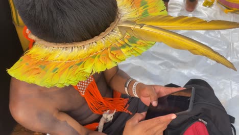 Nahaufnahme-Eines-Jungen-Amazonischen-Mannes-Mit-Traditionellem-Bunten-Federhut,-Der-Sein-Smartphone-Hält