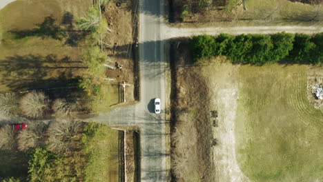 Aerial-shot-of-car-driving-along-rural-country-road-with-trees-and-homes-on-either-side