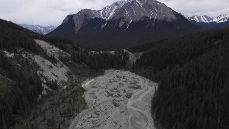 Aerial-view-of-a-dried-creek-surrounded-by-alpine-green-forest-with-a-rocky-mountain-background-with-snow-on-top,-tilt-up-shot,-conservation-concept