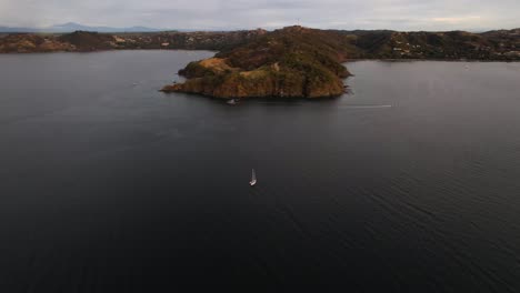 Flying-drone-approaching-single-sailboat-navigating-in-parallel-to-Costa-Rica-rocky-coastline