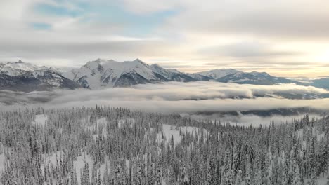 Wunderschöne-Luftaufnahme-Der-Verschneiten-Kanadischen-Rocky-Mountains-Mit-Niedrigen-Wolken-Und-Einem-Grünen-Wald