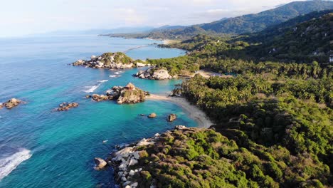 Aerial-view-overlooking-calm,-turquoise-ocean,-beaches-and-the-rocky-shore-of-Tayrona-national-natural-park,-on-a-sunny-evening,-in-the-Caribbean-region-of-Colombia---rising,-drone-shot