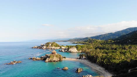 Aerial-view-over-calm,-turquoise-ocean,-beaches-and-the-rocky-coast-of-Tayrona-national-natural-park,-during-golden-hour,-inColombia---pull-back,-drone-shot