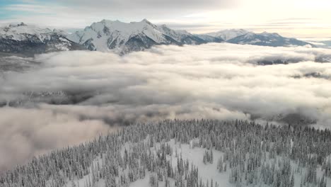 Toma-Aérea-Inclinada-Sobre-Una-Montaña-Cubierta-De-Nieve-Con-Pinos-Y-Nubes-Bajas-En-El-Parque-Nacional-De-Revelstoke,-Canadá