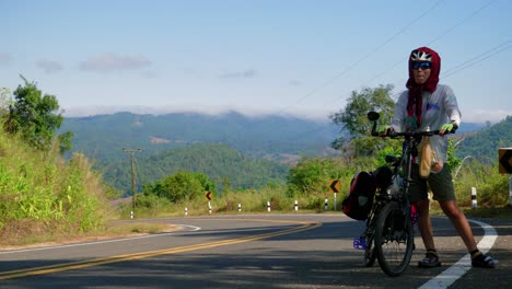 A-lone-female-cyclists-rests-on-a-road-near-Paknai's-Fisherman-Village-before-continuing-by-pushing-her-bicycle-in-the-Na-Muen-District,-Nan,-Thailand