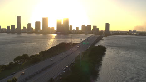Miami-Bridge-Mit-Vorbeifahrenden-Autos-Und-Der-Innenstadt-Von-Miami-Im-Hintergrund-Bei-Sonnenuntergang,-Im-Hintergrund-Scheint-Die-Sonne