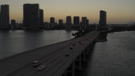 Miami-Bridge-Mit-Vorbeifahrenden-Autos-Und-Der-Innenstadt-Von-Miami-Im-Hintergrund-Bei-Dämmerung.-Luftaufnahme