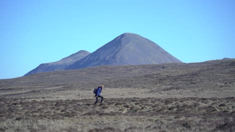 Male-hiking-over-a-mountain-on-a-sunny-day-with-a-high-peak-in-the-background-in-Scotland