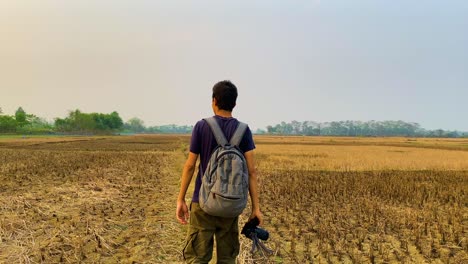 A-young-man-walking-through-a-field-after-harvest-with-a-grey-backpack-and-camera-with-a-tripod-in-his-hand-and-a-forest-in-the-background