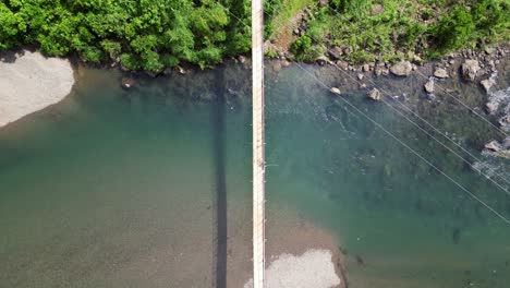 Luftaufnahme-Von-Oben-Nach-Unten-über-Der-Hängebrücke,-Die-Den-Türkisfarbenen-Fluss-Bis-Zum-Grasbewachsenen-Ufer-überspannt
