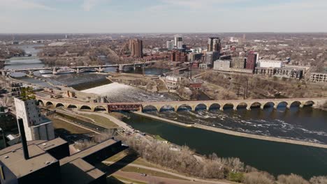 Drone-footage-of-iconic-Stone-Arch-Bridge-over-Mississippi-River-in-Downtown-Minneapolis,-surrounded-by-scenic-cityscape
