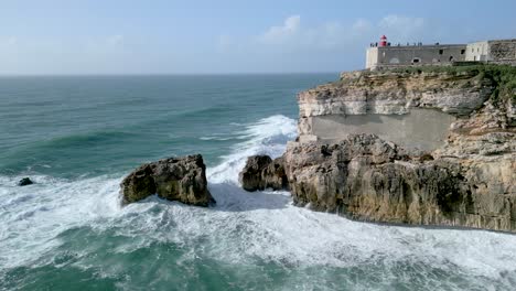 Atemberaubender-Blick-Auf-Den-Leuchtturm-Von-Nazare-Auf-Der-Klippe-Mit-Meereswellen,-Die-An-Der-Felsigen-Küste-In-Portugal-Plätschern