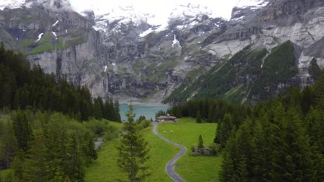 Vista-Aérea-Descendente-Cabaña-De-Madera-Aislada-Chalet-Granja-Solo-En-Verde-Prado-Alpino-Rodeado-De-Montañas-Alpinas,-Pinos-Con-Vistas-Al-Lago-Glaciar-Azul-Turquesa-Oeschinensee-En-Kandersteg,-Suiza