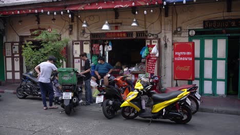 Off-duties-grab-food-rider-and-their-friends-checking-the-bike-conditions-while-the-shop-vendors-look-around-outside-his-shop-in-Yaowarat-Chinatown,-Bangkok,-Thailand