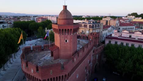 Drone-clip-encircling-Le-Castillet-Fortress-in-Perpignan,-France-overhead-at-sunset,-with-musician-playing-on-rooftop-and-French-flag-flying-in-gentle-breeze