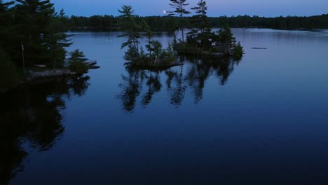 Flying-along-water-surface-over-islands-reflecting-on-Ontario-lake-at-twilight-with-moon-in-sky