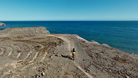 Aerial-shot-of-a-man-with-his-mountain-bike-in-a-desert-landscape-and-where-the-coast-and-the-sea-can-be-seen