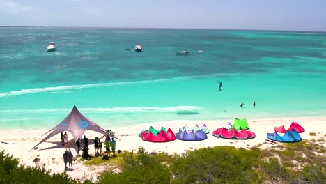 Kitesurfers-cross-and-jump-front-of-caribbean-beach,-aerial-pan-right-crasqui-island