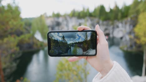 Woman-taking-a-photo-of-a-scenic-nature-view-with-her-phone,-colorful-natural-landscape-with-forest-and-water-pond-in-front-of-her