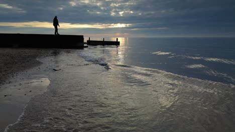 Kleine-Wellen-In-Zeitlupe-Und-Eine-Silhouette-Eines-Mannes,-Der-Bei-Sonnenuntergang-Am-Fleetwood-Beach-In-Lancashire,-Großbritannien,-Zum-Ende-Des-Stegs-Geht